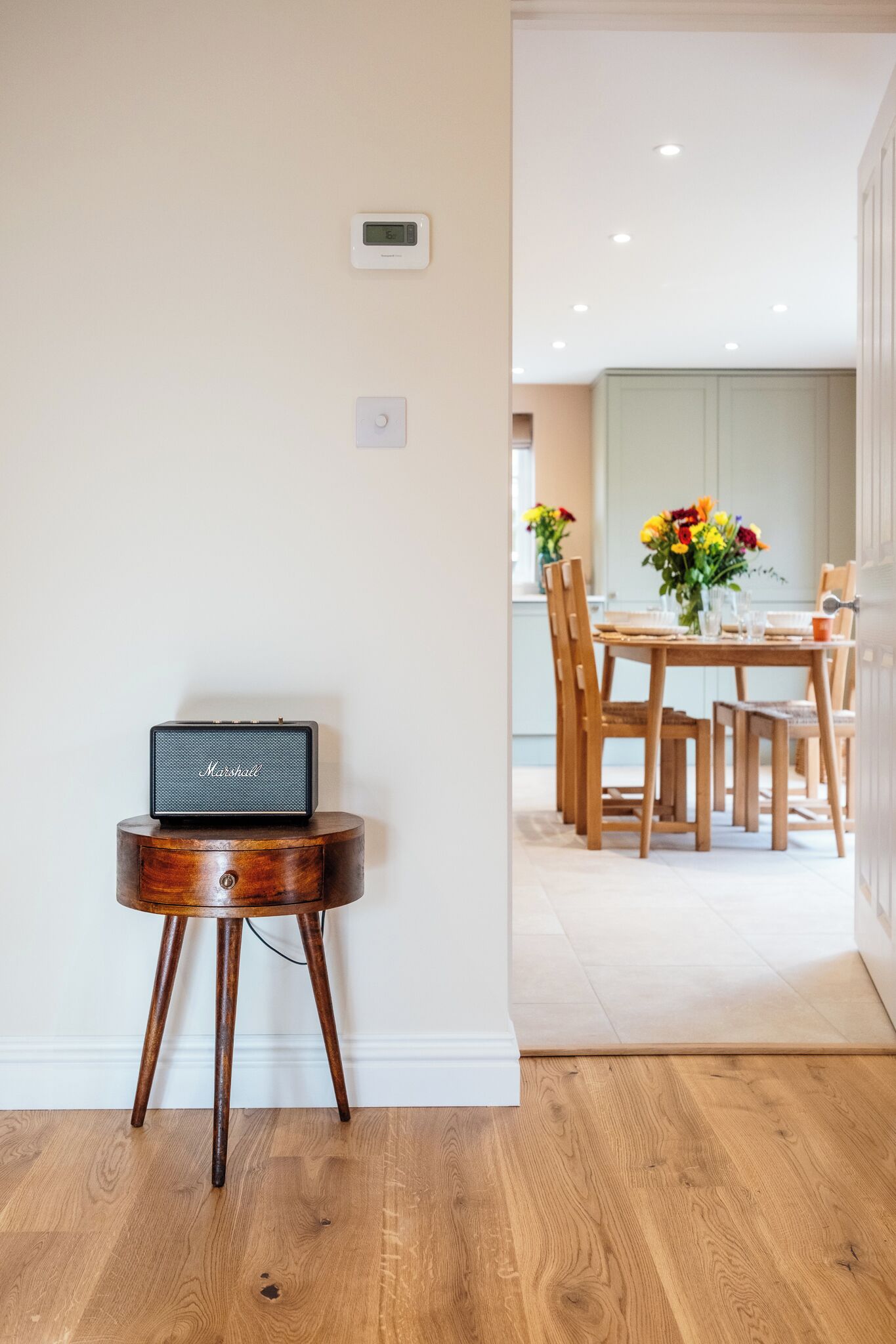 A modern interior with a wooden table and chairs, a small speaker on a wooden stool, and a dining area with a vase of flowers.