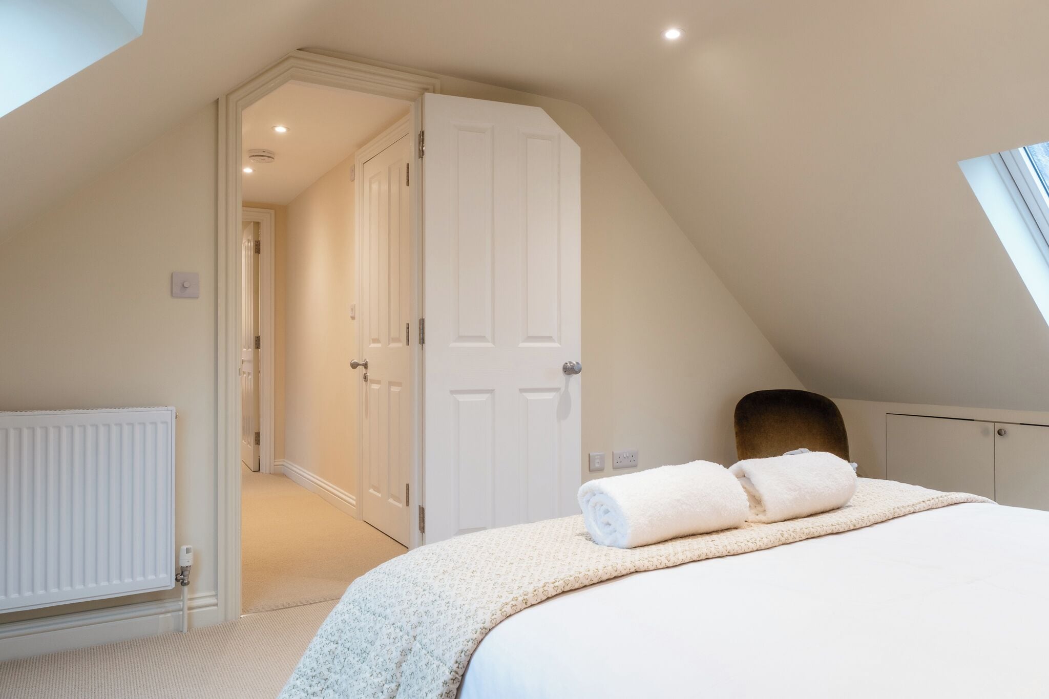 A bedroom with a sloped ceiling, a bed with a white comforter, a radiator, and a door leading to another room.