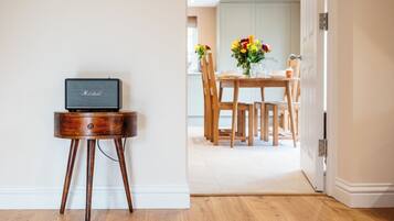 A wooden table with a laptop and a speaker, a dining area with chairs and a table, and a doorway leading to another room.
