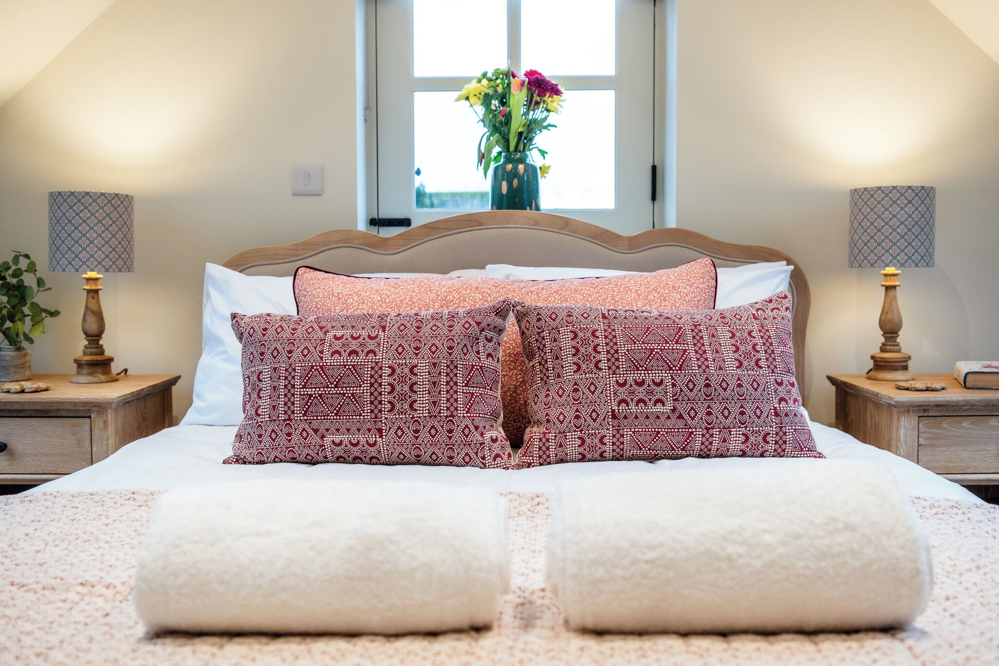 A neatly made bed with patterned pillows, a vase with flowers, and two bedside tables with lamps.