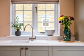 A kitchen with a white sink, a window with a view of buildings, and two vases with flowers. - The Stables Banham Cottages (Bourton-on-the-Water)