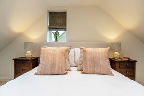A bedroom with a bed, two nightstands, a window with a view, and a patterned window blind. - The Stables Banham Cottages (Bourton-on-the-Water)