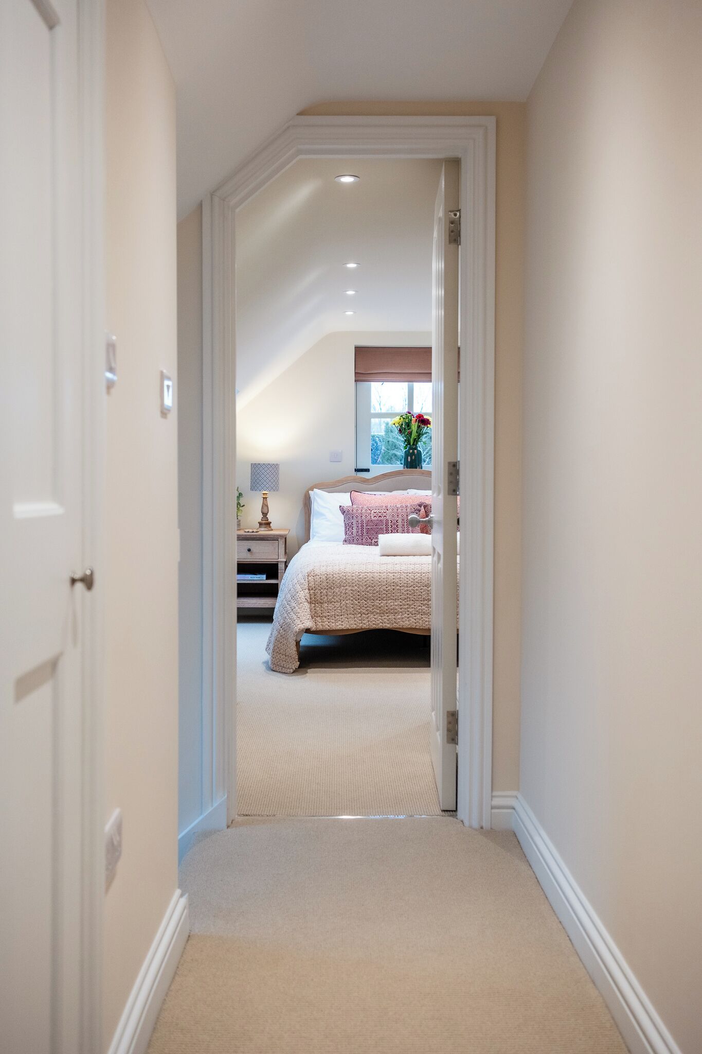 A hallway leading to a bedroom with a bed, bedside table, lamp, and window with flowers.