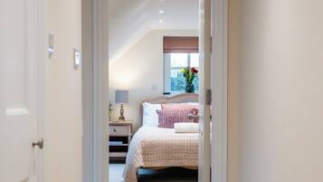 A hallway leading to a bedroom with a bed, bedside table, lamp, and window with flowers.