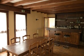 A dining area with a wooden table and chairs, a bar with stools, and shelves with glassware. - Large nature gîte 20 pers - Bar - Veranda - 1 ha - Lake Rillé (Rillé)