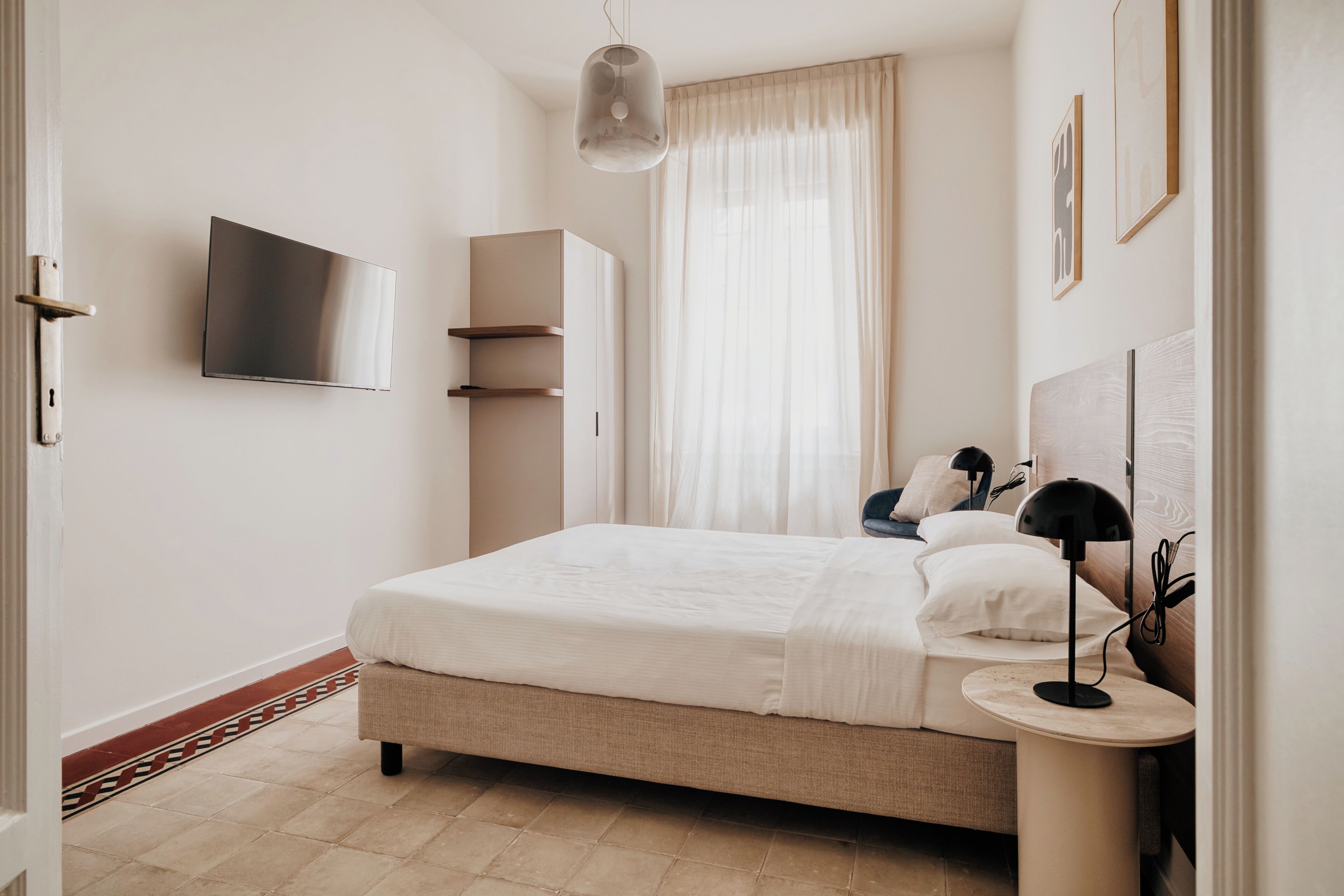 A bedroom with a bed, bedside table, lamp, TV, wardrobe, and a window with curtains.