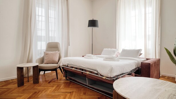 Hotelkamer met een bed, een stoel, een tafeltje, een vloerlamp en een potplant.