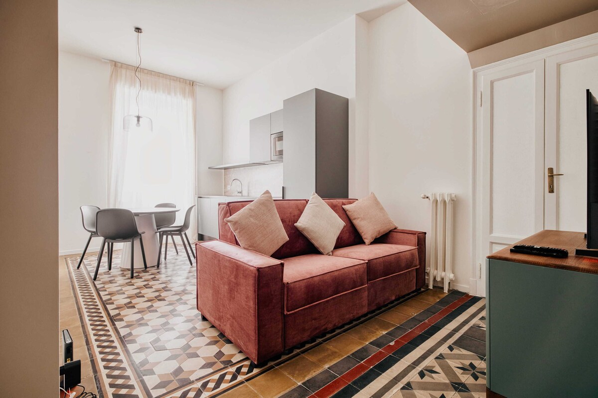 A living room with a red sofa, a dining table with chairs, a kitchen area, and a patterned floor.