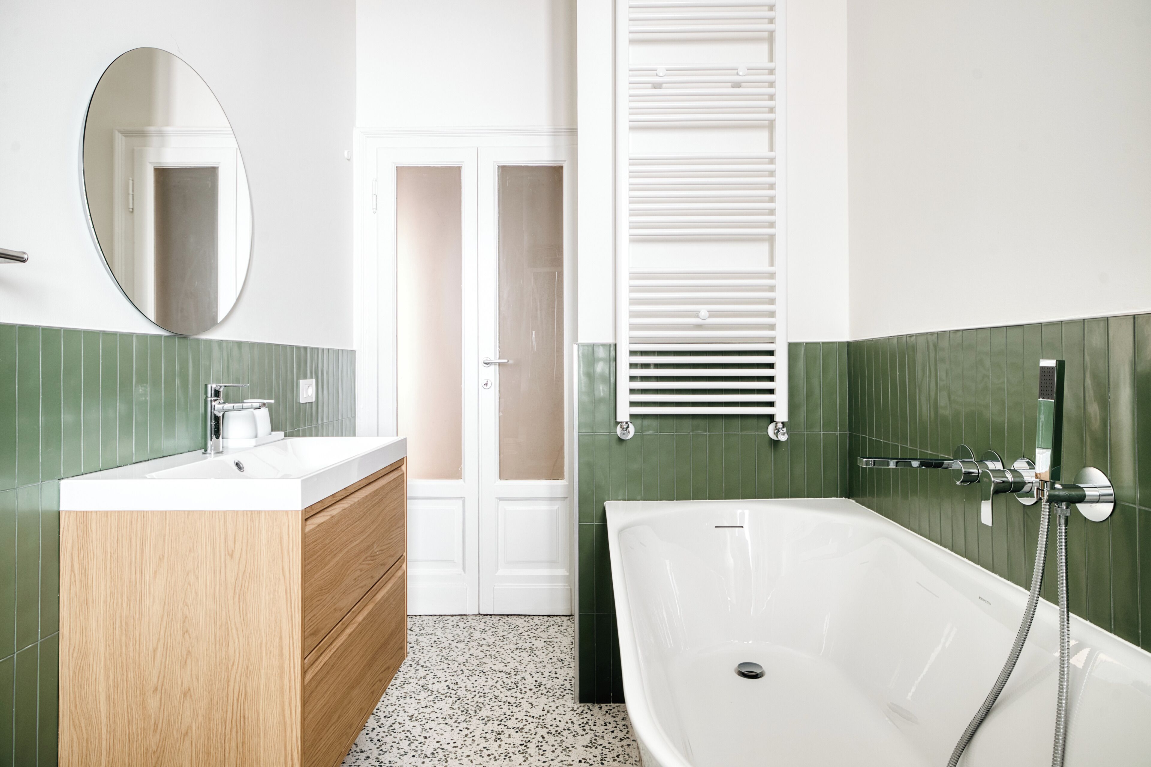 A bathroom with a white bathtub, a wooden vanity, a round mirror, and green tiled walls.