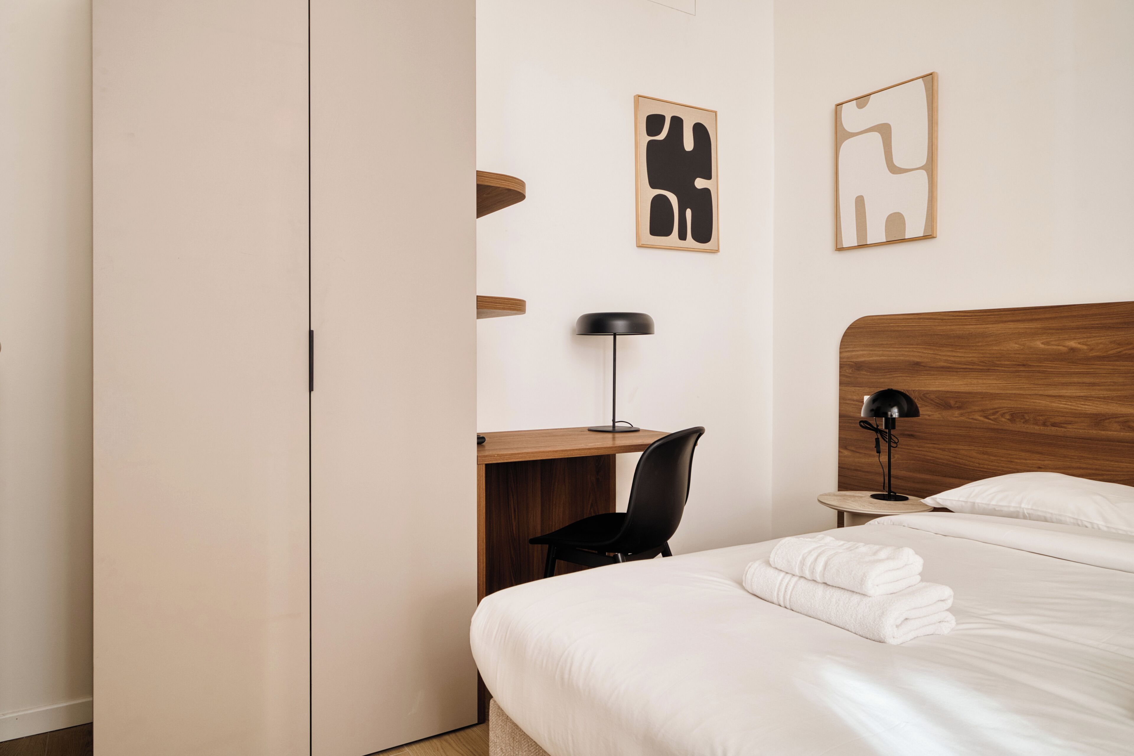 Hotellihuone, jossa on sänky, työpöytä, tuoli, lamppu ja kaksi seinätaideteosta.