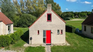 Ein kleines Steinhaus mit roter Tür und Fensterläden, umgeben von Grünpflanzen und weiteren Gebäuden.