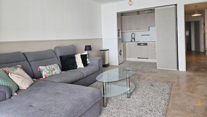 A modern living room with a grey sectional sofa, a glass coffee table, and a kitchen area with light wood cabinets and a washing machine. - Central Juan-les-Pins Colombier 1-Bedroom with Sea View, Parking (Antibes)