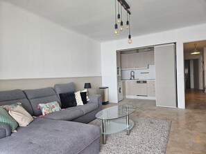 Una sala de estar moderna con un sofá seccional gris, una mesa de centro de vidrio y una zona de cocina con armarios de madera clara y una lavadora.