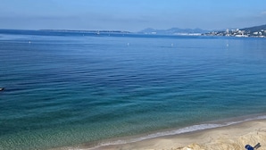 A coastal view with clear water, a sandy beach, and distant buildings. - Central Juan-les-Pins Colombier 1-Bedroom with Sea View, Parking (Antibes)
