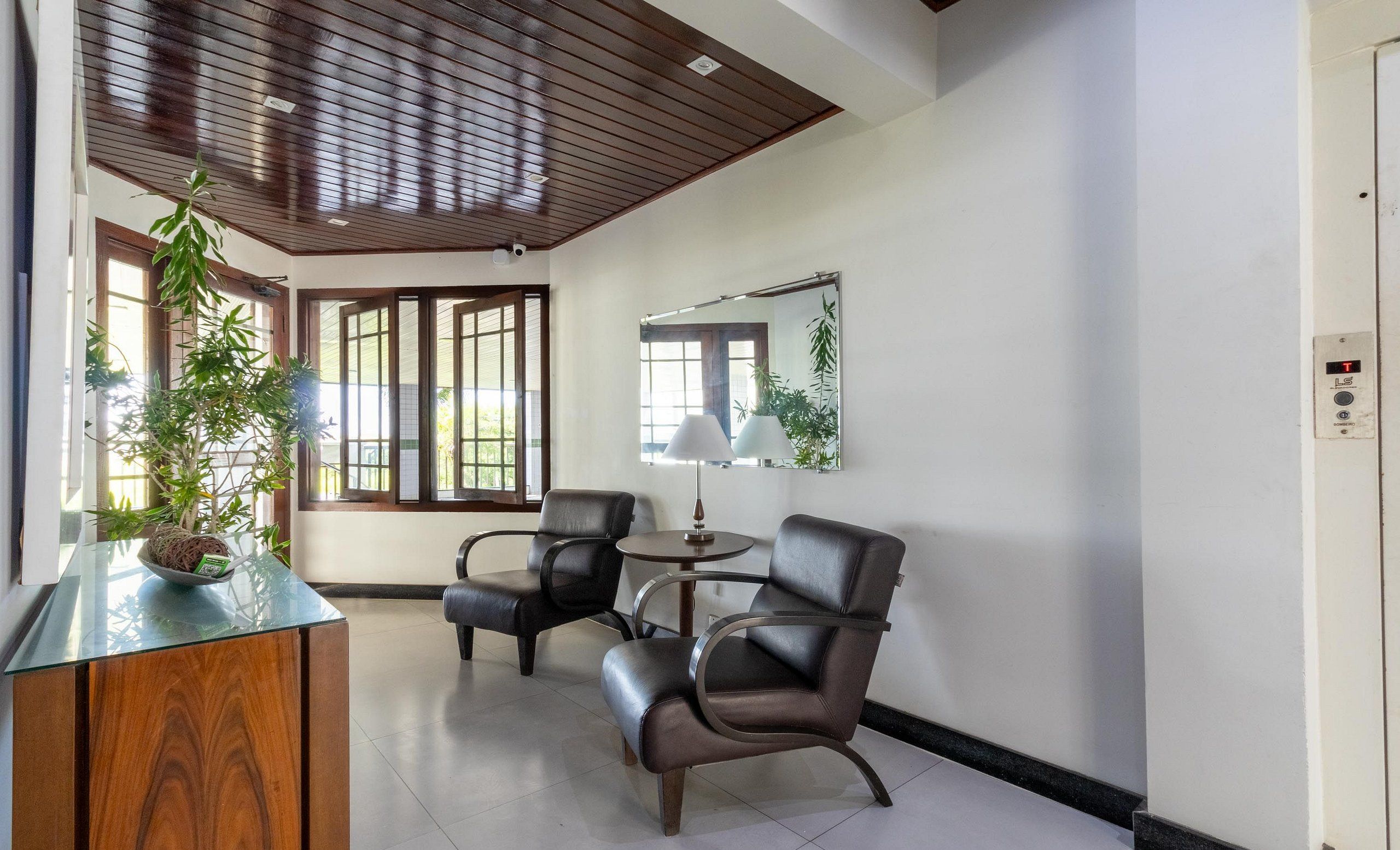 A room with a wooden ceiling, a glass-top desk, two leather chairs, a round table with a lamp, and a large mirror on the wall.