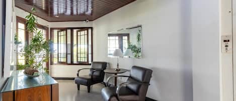 A room with a wooden ceiling, a glass-top desk, two leather chairs, a round table with a lamp, and a large mirror on the wall.