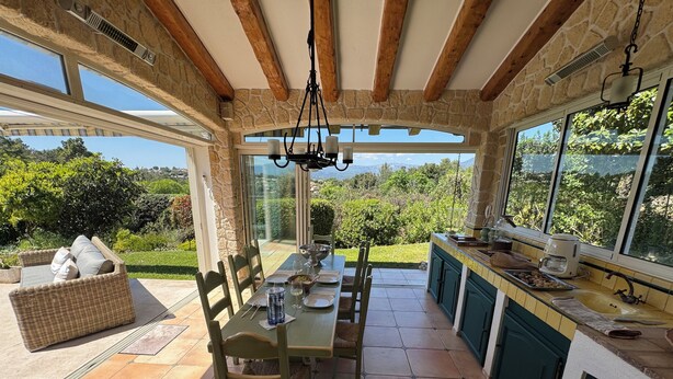 A kitchen with a dining area, stone walls, wooden beams, and large windows overlooking a garden. - Beautiful Villa in private estate with 5 bedrooms, swimming pool, panoramic view (Villeneuve-Loubet)
