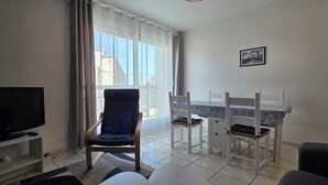 A living room with a TV, a chair, a dining table with chairs, and a window with curtains. - Beautiful, bright, renovated apartment in the center of Concarneau with private parking (Concarneau)