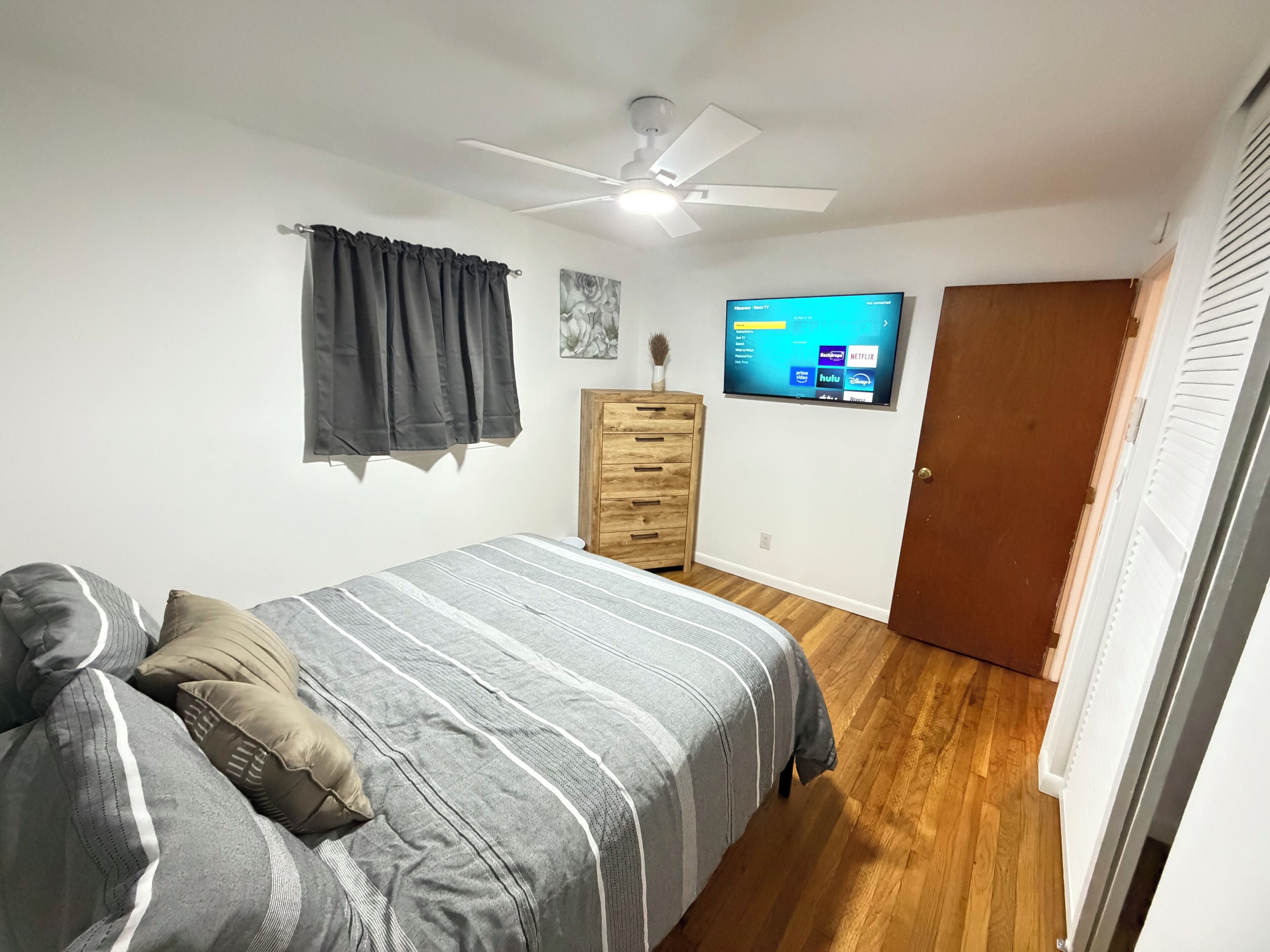 A bedroom with a bed, a dresser, a ceiling fan, a TV mounted on the wall, and a door.