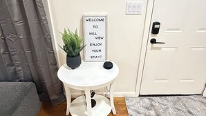 A room with a white door, a welcome sign, a small table with a potted plant, and a grey rug.