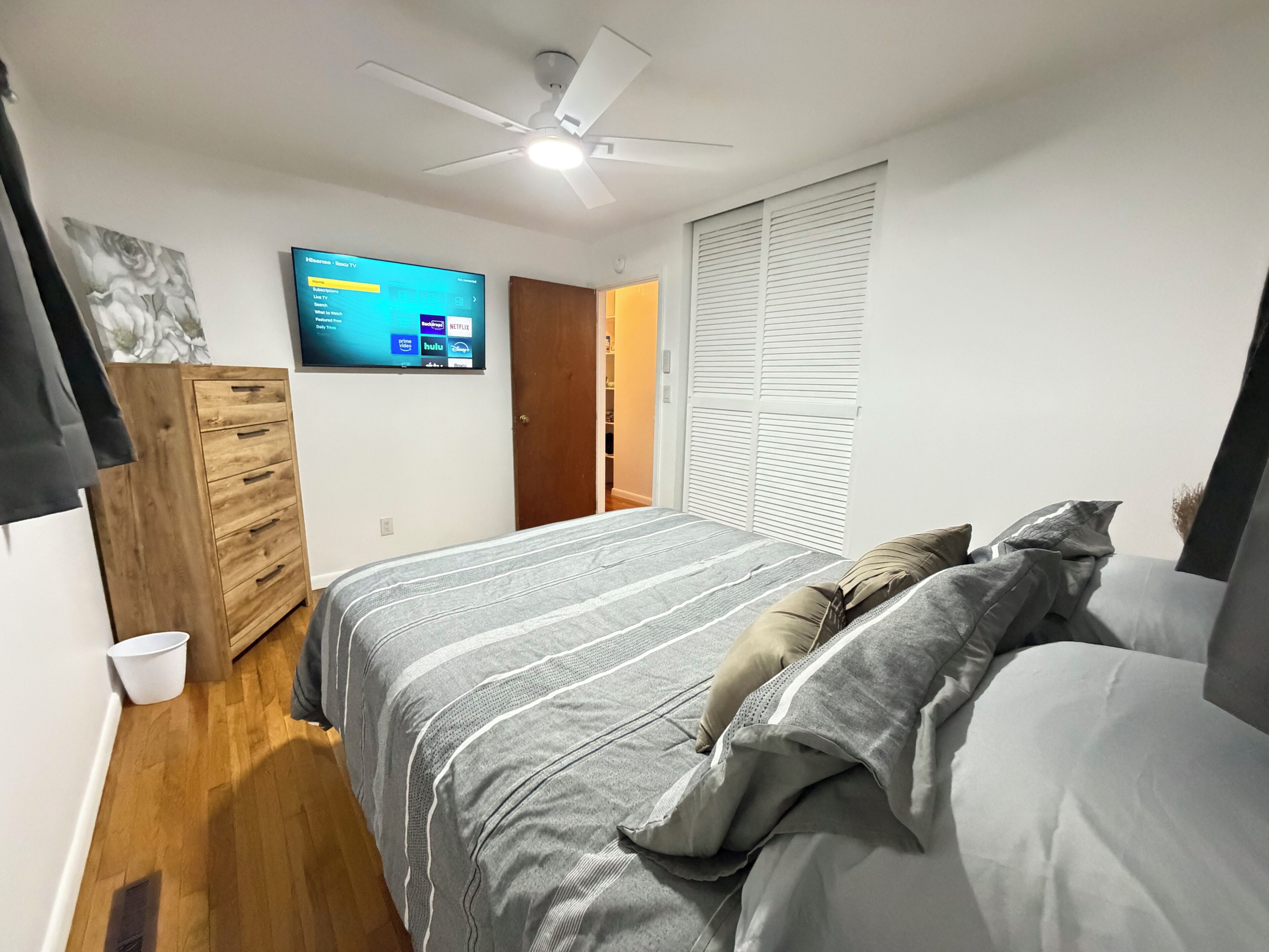 A bedroom with a bed, a ceiling fan, a TV mounted on the wall, a wooden wardrobe, and a white bucket on the floor.