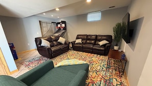 A living room with a sofa, two armchairs, a coffee table, a TV, a basketball hoop, and a patterned rug.