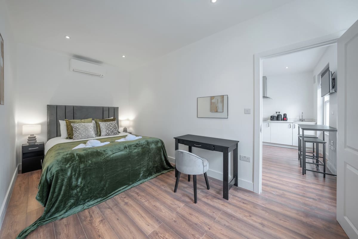 A bedroom with a bed, bedside table, desk, and chair, and a connected kitchen area.