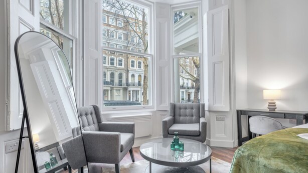 Una habitación con un espejo grande, dos sillones grises, una mesa de centro redonda de vidrio y vistas a edificios a través de amplios ventanales.