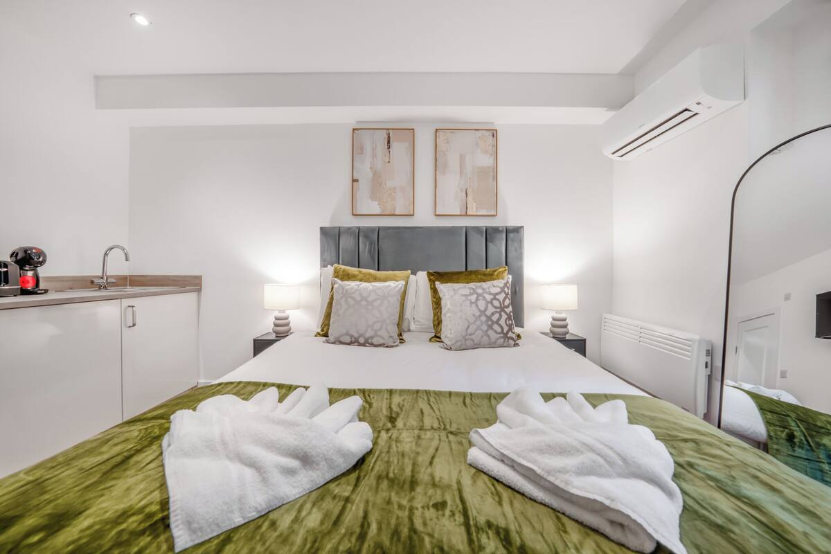 A hotel room with a bed, two bedside tables, a nightstand, a mirror, an air conditioning unit, and a small kitchenette area.