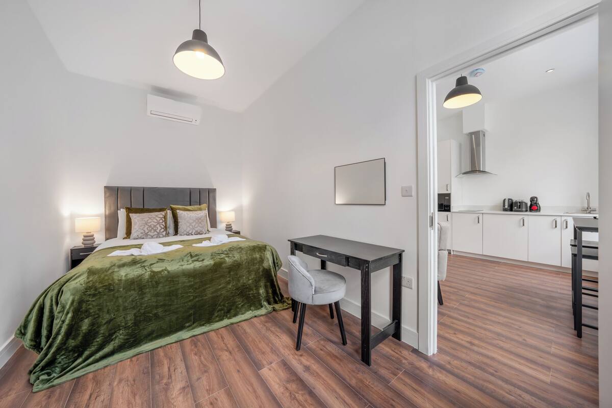 A bedroom with a bed, bedside table, lamp, and a view into the kitchen.