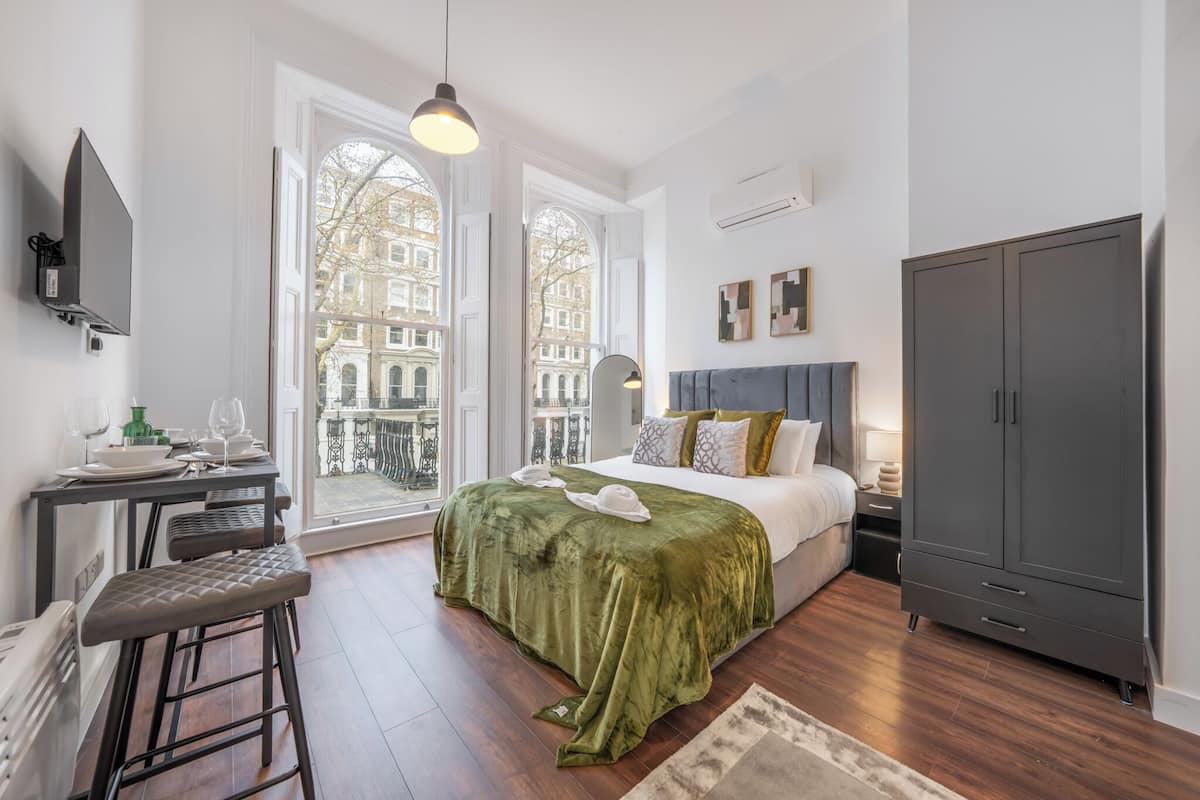 A bedroom with a bed, a desk with a chair, a wardrobe, and large windows with a view of buildings.