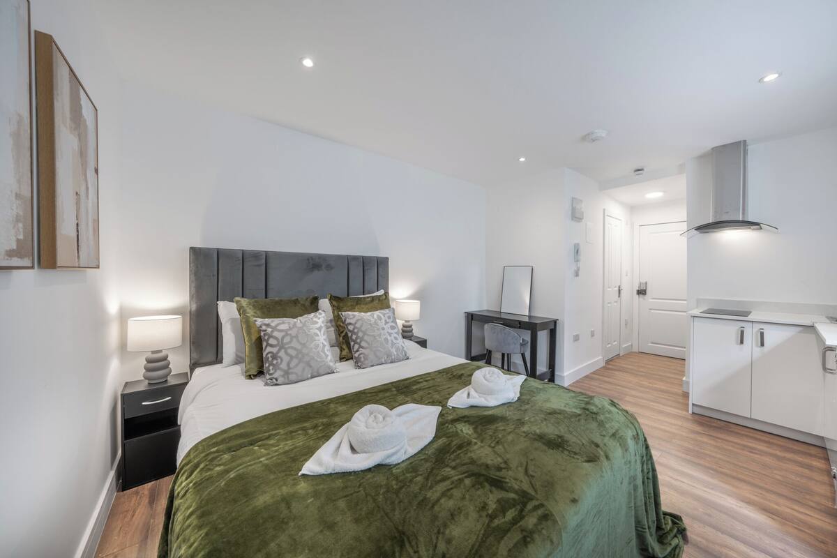 A modern hotel room with a large bed, bedside tables, a desk, and a kitchenette.