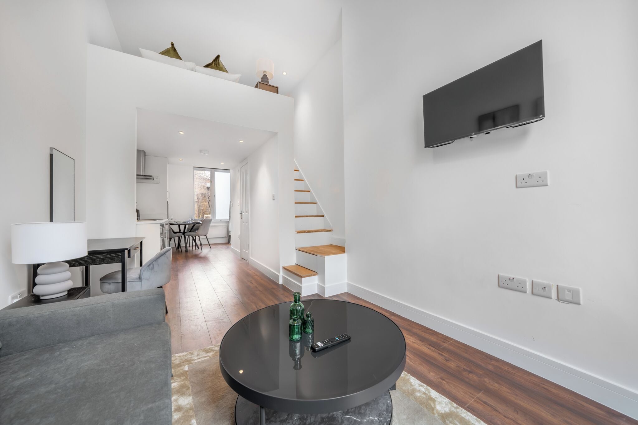 A modern living room with a round black table, a grey sofa, a TV mounted on the wall, and a staircase leading to an upper level.