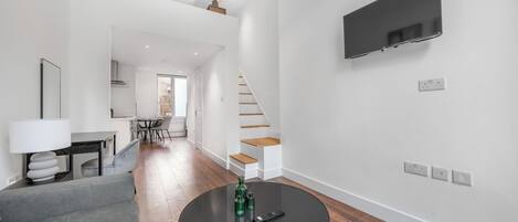 A modern living room with a round black table, a grey sofa, a TV mounted on the wall, and a staircase leading to an upper level.