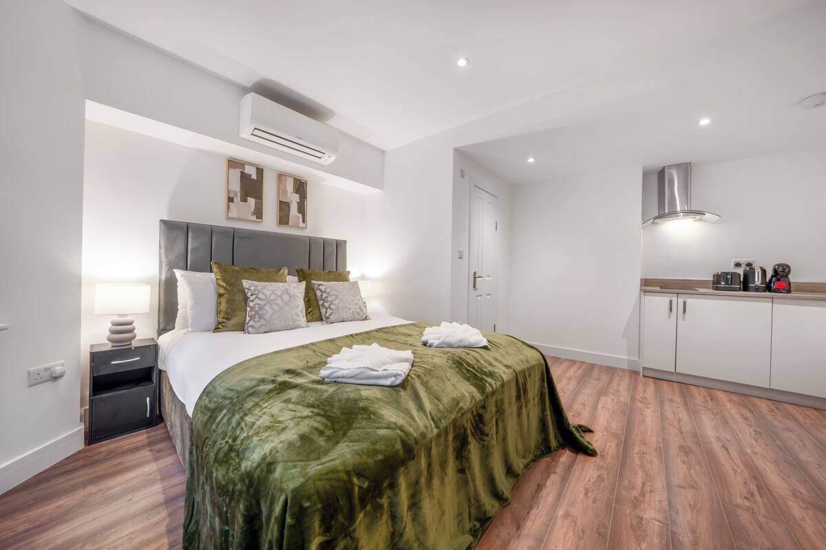 A modern hotel room with a large bed, a bedside table, a built-in wardrobe, and a kitchenette area.