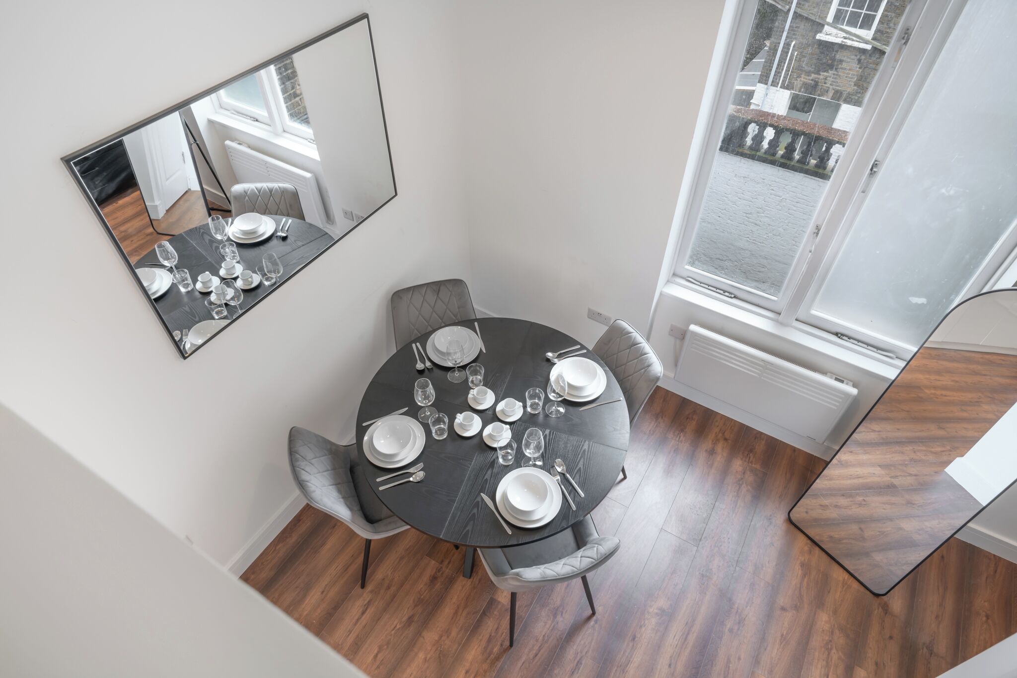 Un comedor con una mesa redonda dispuesta para cuatro, un espejo en la pared y vistas al exterior a través de una ventana.