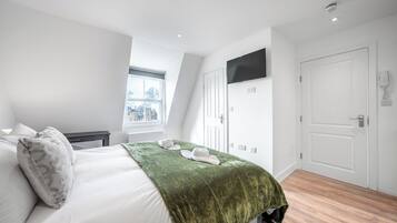A hotel room with a bed, a green throw, a flat-screen TV, a white door, and a window with a city view.