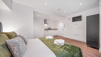 A hotel room with a bed, a green bedspread, a TV, a wardrobe, and a kitchenette in the background.