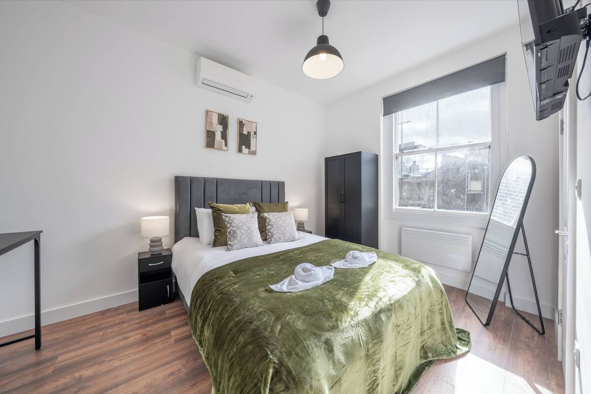 A modern bedroom with a large bed, bedside tables, a standing mirror, and a window with a view.