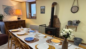 Un coin repas avec une longue table dressée pour six personnes, une cheminée en briques et une horloge au mur.