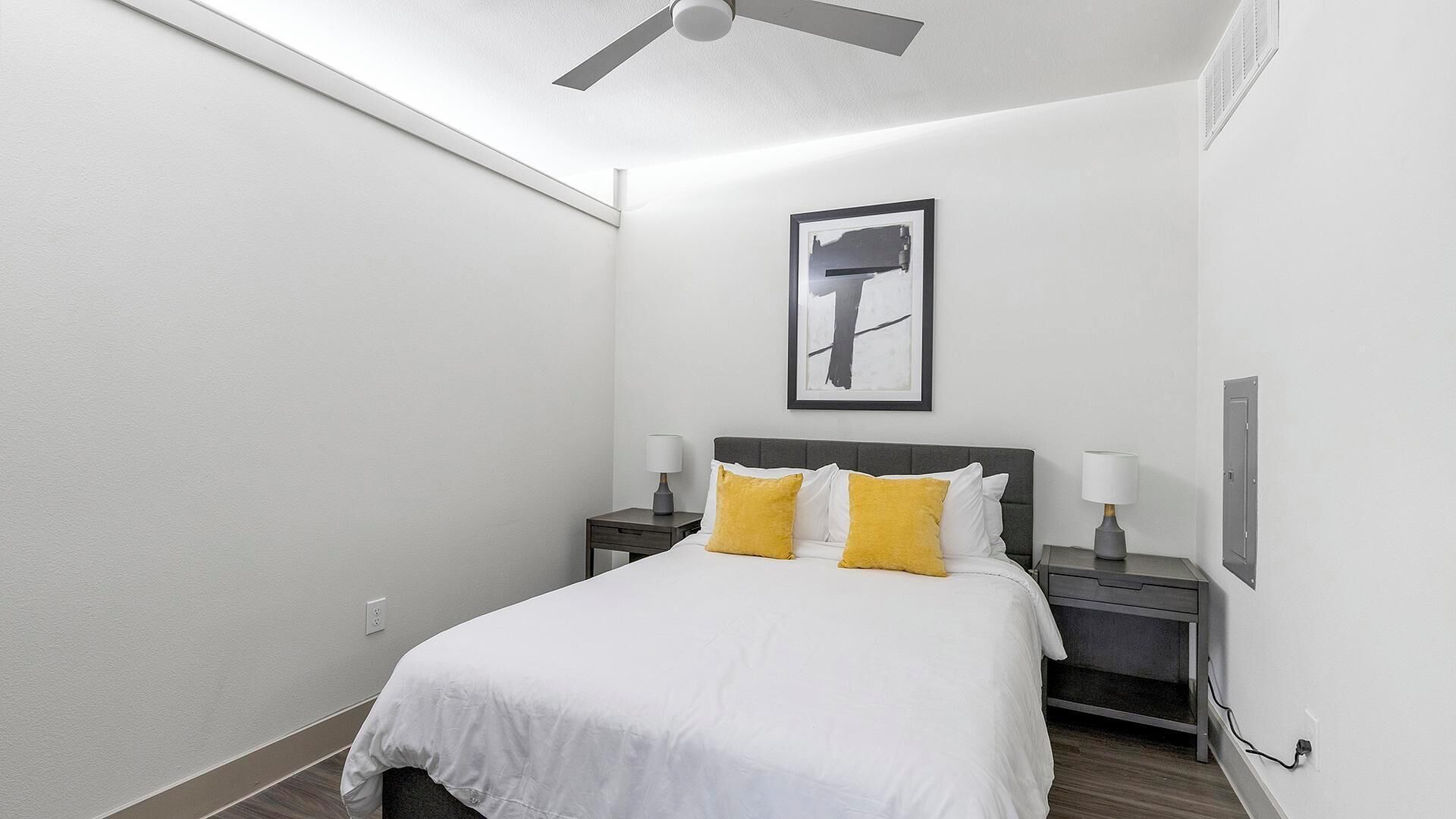 A hotel room with a bed, two bedside tables, a ceiling fan, a framed picture on the wall, and a power outlet on the wall.