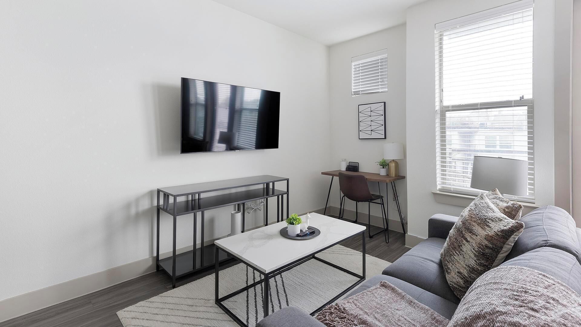 Een moderne woonkamer met een flatscreen televisie aan de muur, een eethoek met een tafel en stoelen, en een bank met kussens.