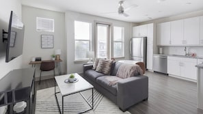 Un apartamento moderno con un sofá gris, una mesa de comedor, una cocina con armarios blancos y un ventilador de techo.
