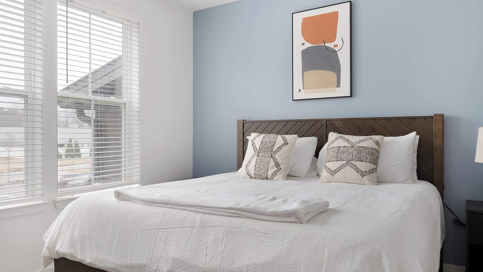 Un dormitorio con cama, almohadas, cabecero, ventana con persianas y una obra de arte colgada en la pared.