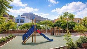 Un parque infantil con tobogán, estructuras para escalar y vistas a edificios y montañas al fondo.