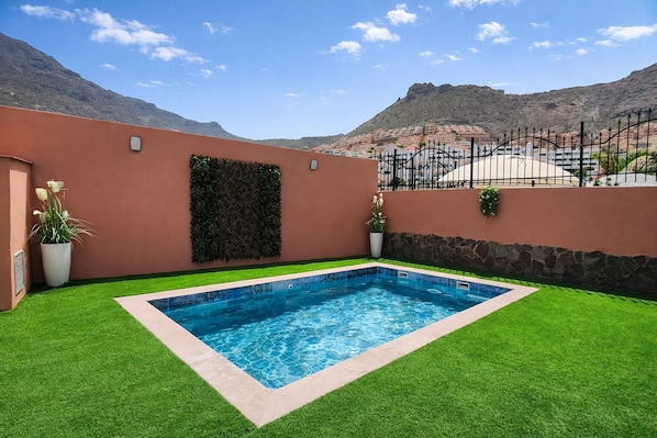 Une piscine rectangulaire entourée de gazon synthétique, un mur de pierre et une vue sur un paysage montagneux.