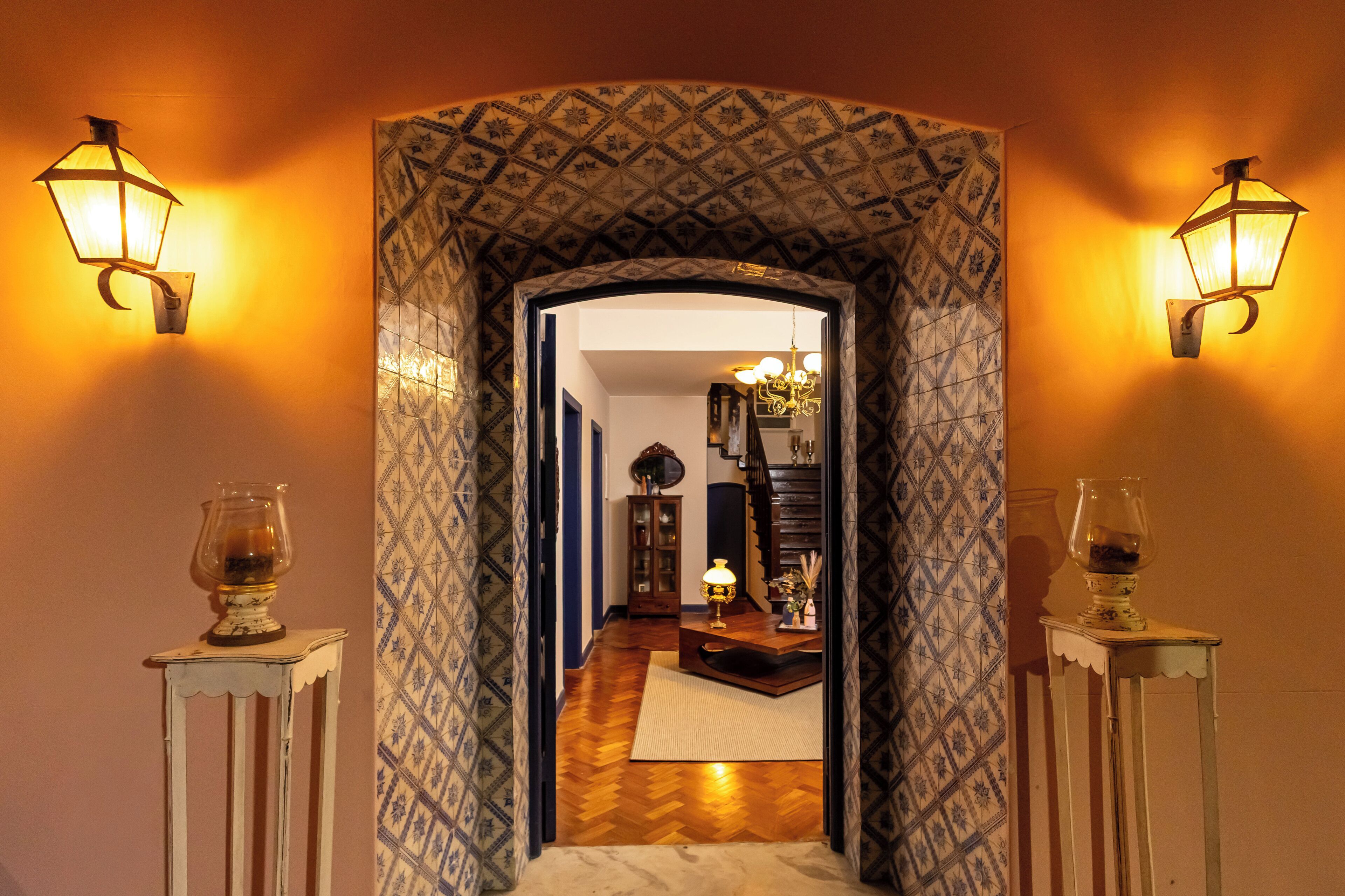 A hallway with patterned wallpaper, two wall-mounted lamps, and a mirror reflecting a room with a chandelier.