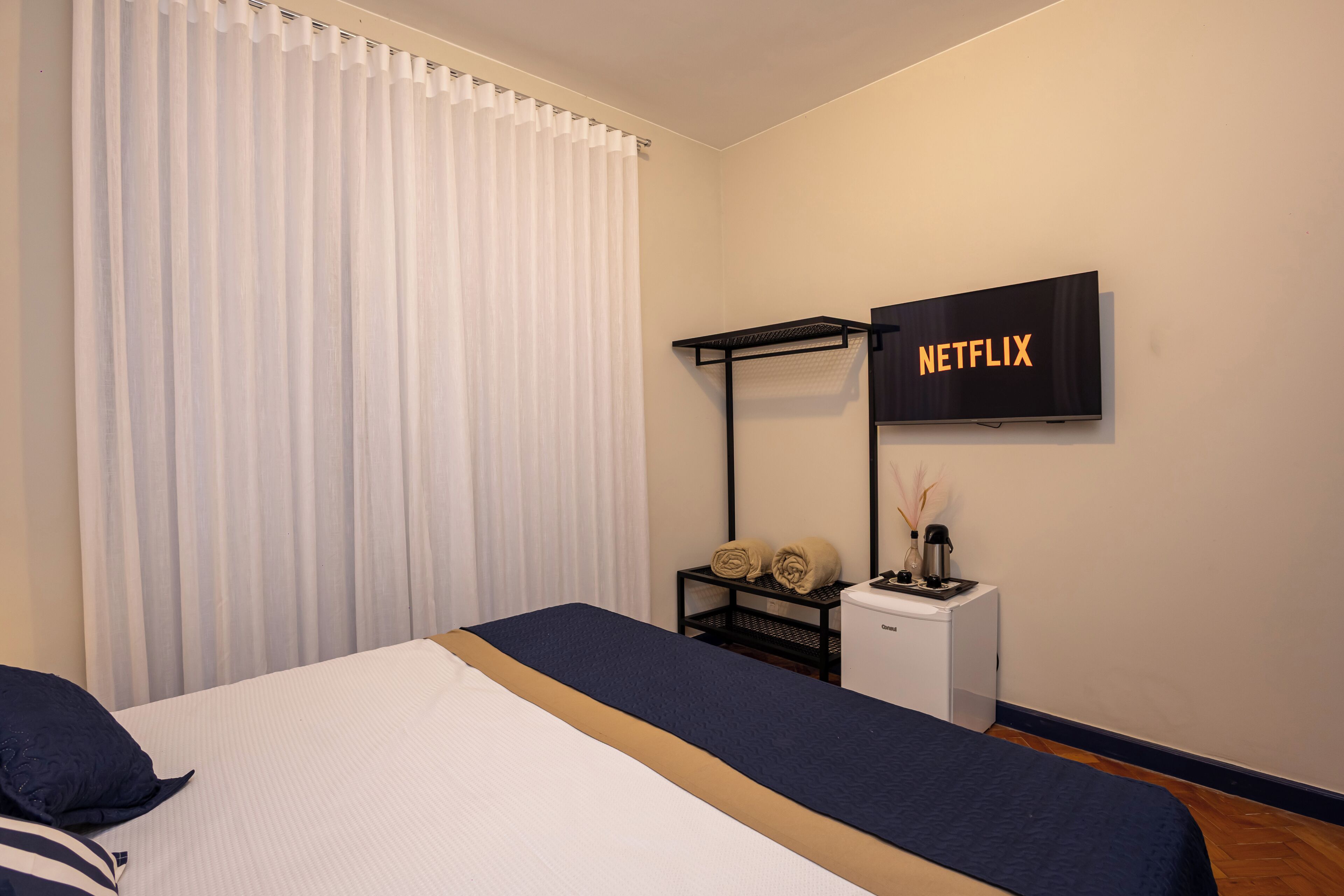 A hotel room with a bed, a TV mounted on the wall, a small desk with a lamp, and a curtain.