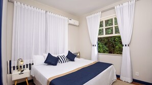 A neatly arranged hotel room with a bed, curtains, an air conditioning unit, a window with a view, and a bedside table with a lamp.