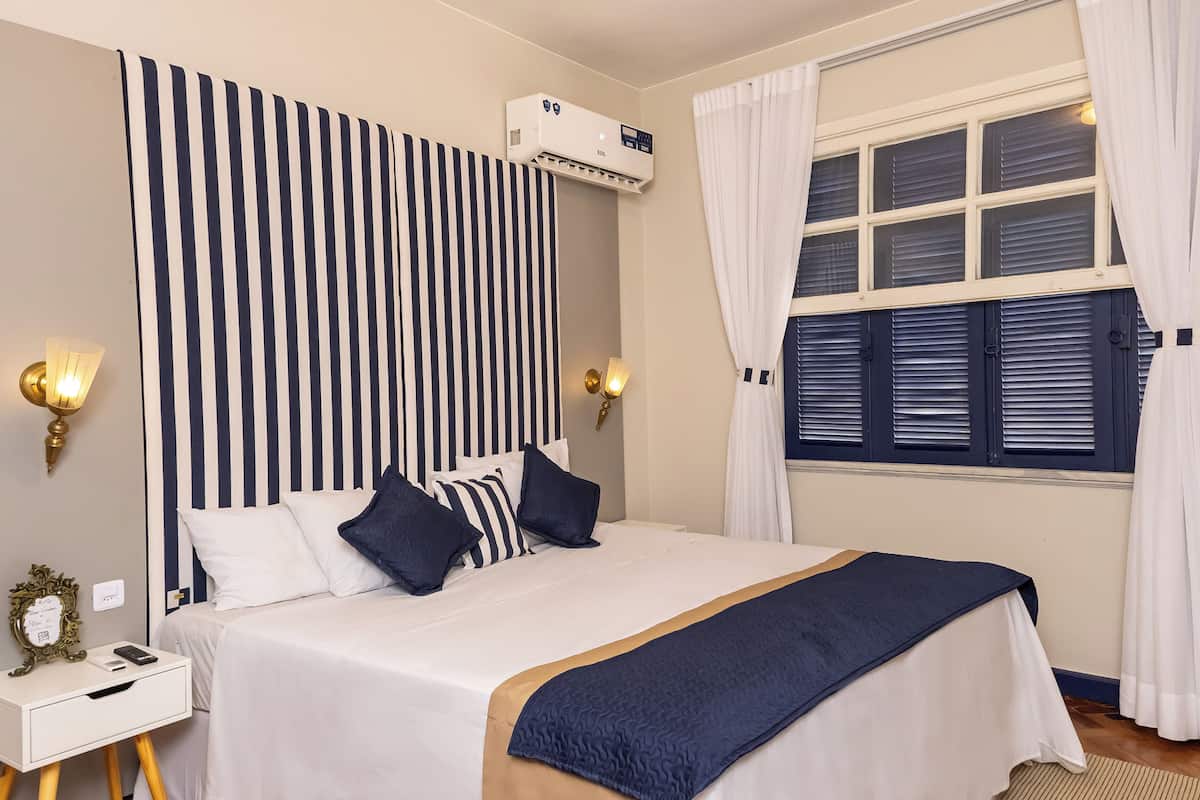 A hotel room with a bed, bedside lamps, a nightstand, a window with curtains, and an air conditioning unit.